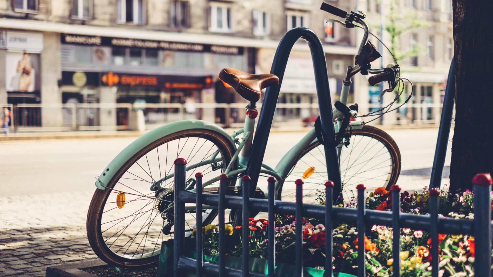 Ein Fahrrad lehnt leicht schräg gegen einen Metallbügel. Im Hintergrund sind Geschäfte zu sehen.